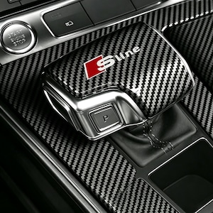May include: A car gear shift knob cover featuring a black and gray carbon fiber pattern with a red and white S-line logo. The cover includes a metallic silver finish on the side with a button labeled P, positioned within a carbon fiber console.
