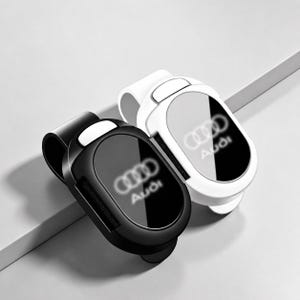 Peut inclure: Deux accessoires d'intérieur de voiture, l'un en noir mat et l'autre en blanc brillant, chacun présentant une face ovale lisse avec le logo et le texte Audi imprimés au centre. Ces clips fonctionnels ont une forme incurvée pour les aérateurs.