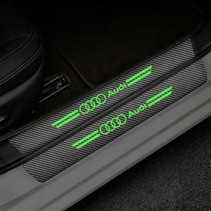May include: Two rectangular carbon fiber textured door sill protectors for a vehicle. Each protector features the Audi logo and brand name in a bright, luminous green color, accompanied by two horizontal green lines on either side of the text.