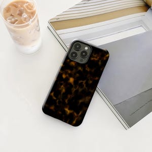 May include: A tortoiseshell-patterned phone case with a glossy finish. The case is dark brown with amber and black marbling. The phone case is lying on a white surface next to a glass of iced coffee and a magazine.
