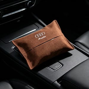 Peut inclure: Un porte-mouchoirs en suédine marron arborant le logo et le texte Audi brodés en blanc sur le devant. Cet accessoire rectangulaire présente une fente horizontale centrale pour distribuer les mouchoirs et est conçu pour l'intérieur d'un véhicule.