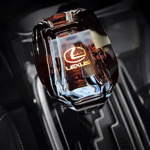 May include: A translucent, dark amber-coloured gear shift knob with a faceted, crystal-like design. The Lexus logo and the word LEXUS are clearly visible in gold lettering inside the centre of the knob, which is mounted on a black gear shifter base.