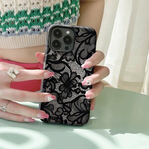May include: Black phone case with a detailed lace pattern. The case features a floral design and a camera cutout. The case is held by a person with long, manicured nails. The case is a sleek accessory.