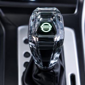 May include: A clear, faceted crystal gear shift knob featuring a glowing green Nissan logo at the top. The gear shifter has a geometric, multi-faceted design and is attached to a black leather boot, positioned within a vehicle centre console.