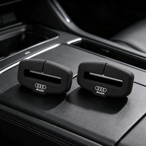 Może przedstawiać: Dwie matowe czarne osłony na klamry pasów bezpieczeństwa z białym logo Audi i emblematem czterech pierścieni. Te prostokątne silikonowe akcesoria posiadają centralne poziome wycięcie oraz pionową szczelinę, pasujące do standardowych klamer.