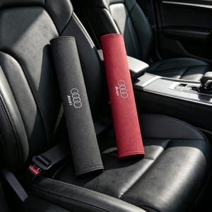May include: Two rectangular car seat belt covers, one in black and one in red, featuring a perforated texture and the white Audi logo embroidered on the front. These protective pads are designed to wrap around standard vehicle seat belt straps.