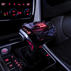 May include: A black gear shift knob for an Audi vehicle featuring a glossy, reflective top surface with the illuminated red Audi logo. The side of the knob displays the gear positions P, R, N, and D/S in red light, set within a modern car interior.