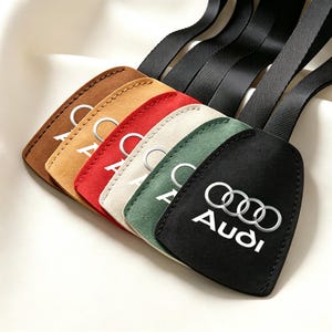 May include: A set of five suede-like key tags in brown, tan, red, cream, and black, each featuring the white Audi logo and text. Each tag is attached to a black fabric strap, arranged in a fanned-out display on a soft, off-white fabric surface.