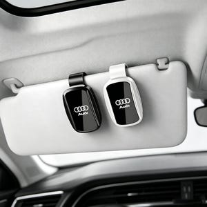May include: Two car visor clips attached to a light gray vehicle sun visor. One clip is black and the other is white. Both clips feature a glossy black center panel with the silver Audi logo and the word Audi printed in white text on the surface.