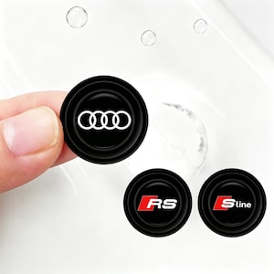May include: Three circular black emblems with white and red detailing. One features the white Audi four-ring logo, another displays the red and white RS emblem, and the third shows the red and white S-line logo. Each emblem has a smooth, rounded edge.