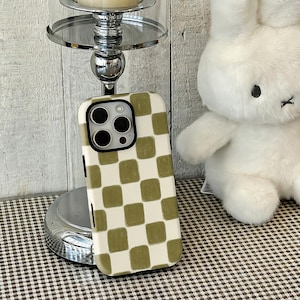 Puede incluir: Una funda de teléfono a cuadros blancos y verde oliva. La funda tiene un recorte para la cámara y bordes negros. Un peluche de conejo blanco está en el fondo, junto con un candelabro plateado.