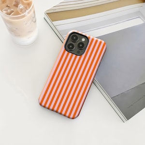 May include: A smartphone case featuring a vertical striped design in orange and pale pink. The case is placed on a white surface, alongside a magazine and a glass of iced coffee.