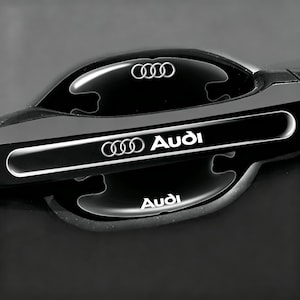 May include: A black car door handle cover featuring the silver Audi logo and text. The design includes a horizontal strip with the Audi emblem and brand name, plus a secondary Audi logo and text positioned on the lower section of the handle.