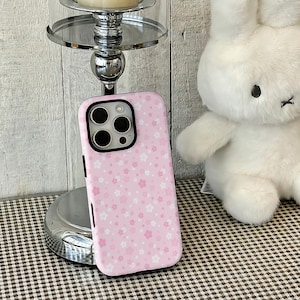 Puede incluir: Una funda de teléfono rosa con un estampado floral. La funda tiene un recorte negro para la cámara y botones laterales negros. Descansa sobre una superficie a cuadros, junto a un conejito de peluche blanco y un candelabro plateado.