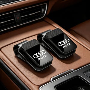 Puede incluir: Dos clips negros para gafas de sol en el parasol del coche con acabado brillante y el logotipo blanco de Audi con cuatro anillos entrelazados. Cada clip tiene una sección superior texturizada y un diseño curvo y robusto para el parasol.