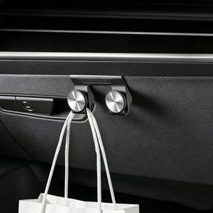 May include: A matte black car interior hook featuring two circular silver metallic knobs with a radial brushed finish. The hook is mounted on the dashboard and holds the white cord handles of a shopping bag, providing a storage solution.