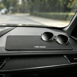 Könnte beinhalten: Eine rechteckige schwarze Armaturenbrettmatte mit horizontaler Rippenstruktur und dem in Weiß aufgedruckten Audi-Logo. Die Matte liegt auf dem Armaturenbrett des Autos, darauf befindet sich eine silberne Sonnenbrille mit dunklen Gläsern.