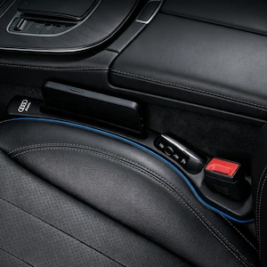 Puede incluir: Un organizador de huecos para asientos de coche en color negro con un perfil delgado, acabado mate liso y una tira de luz LED azul a lo largo del borde. La unidad incluye una ranura para un smartphone y muestra el logotipo de Audi en blanco.