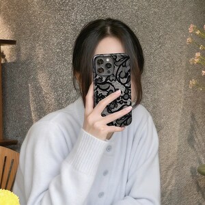 May include: Black and gray phone case with a delicate lace pattern. The case has a glossy finish and a dark border. The design features floral and swirling elements, creating an elegant and intricate look. The case is shown being held by a person.