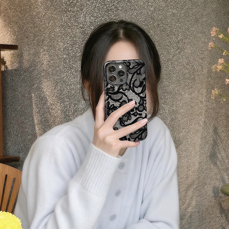 May include: Black and gray phone case with a delicate lace pattern. The case has a glossy finish and a dark border. The design features floral and swirling elements, creating an elegant and intricate look. The case is shown being held by a person.
