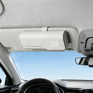 May include: A light gray rectangular car visor sunglasses holder with a magnetic closure, featuring the white Audi logo and text printed on the front. The case is securely attached to the vehicle's sun visor above the steering wheel.