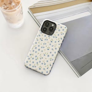 May include: A white phone case with a floral pattern of small blue flowers and green stems. The case is on a white surface next to a magazine and a glass of iced coffee.