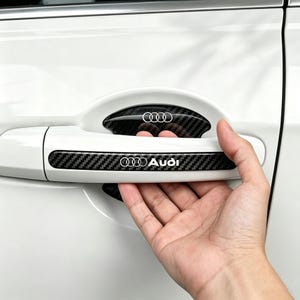 May include: A white car door handle featuring a black carbon fiber textured trim piece with the Audi logo and text. The trim is applied to the handle and the recessed area behind it, showcasing a repeating weave pattern and silver branding.