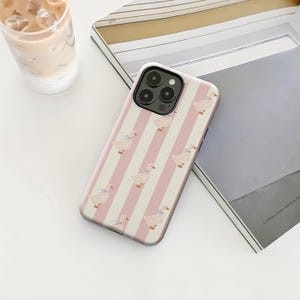 May include: A phone case featuring a pattern of pink and white vertical stripes with repeating illustrations of ducks wearing blue bows. The case is designed to fit a smartphone and has a black camera lens cutout.