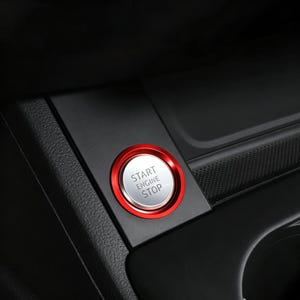 May include: A circular car engine start stop button featuring a metallic silver centre with the text START ENGINE STOP printed in black, surrounded by a vibrant red outer ring, mounted within a black textured plastic console panel.