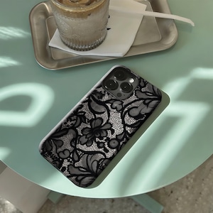 May include: A black and white lace patterned phone case. The case features a detailed floral design with intricate patterns. The phone case is on a light green table, with a drink and a tray in the background.