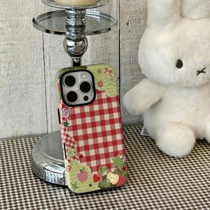 Può includere: Una custodia per telefono decorativa con un motivo a quadri rossi e bianchi, accentuata da disegni floreali e fruttati verdi e rosa. La custodia è su un supporto argentato, con un coniglietto di peluche bianco sullo sfondo.