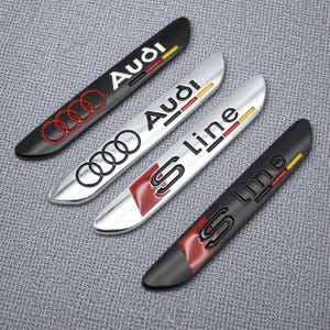 Può includere: Quattro emblemi automobilistici allungati e curvi. Due sono neri e due sono argentati. Due presentano il logo e il testo Audi, mentre due presentano il testo S line. Ogni emblema include una piccola striscia rossa e gialla all'estremità.
