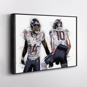 May include: A framed art print featuring two football players in action. The players are wearing white and blue uniforms with the numbers 14 and 10. The artwork has a watercolor effect with splashes of color and a white background.