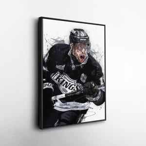 May include: Framed art print of a Los Angeles Kings hockey player in action. The player is wearing a black and white uniform with the team's logo. The artwork has a watercolor-like effect with paint splatters in the background.