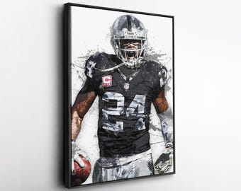 May include: Framed artwork featuring a football player in a Raiders uniform, number 24. The player is in a dynamic pose, holding a football. The artwork uses a watercolor style with black, white, and silver tones.