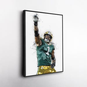 May include: Framed art print of a football player in a green and gold uniform, raising his arm in a gesture of triumph. The artwork features a watercolor-style effect with paint splatters, set against a white background. The player wears a helmet and the number 1.