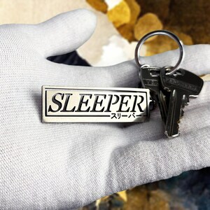 Pode incluir: Chaveiro retangular prateado com a palavra "SLEEPER" em letras maiúsculas pretas. Abaixo está o texto japonês. O chaveiro está preso a um chaveiro prateado com duas chaves.
