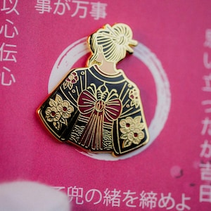 Może przedstawiać: Złoty emaliowany pin przedstawiający kobietę w tradycyjnym japońskim kimono. Czarny kimono zdobiony jest złotymi kwiatowymi i geometrycznymi wzorami, dużą czerwoną kokardą i czerwoną frędzlą. Pin umieszczony jest na różowym tle z japońskim tekstem.