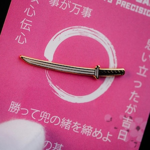 Könnte beinhalten: Goldfarbene Katana-Anstecknadel mit schwarzen und weißen Details, vor einem rosa Hintergrund mit japanischem Text und einem weißen Kreisdesign. Die Anstecknadel ist eine Miniaturreplik eines traditionellen japanischen Schwertes.