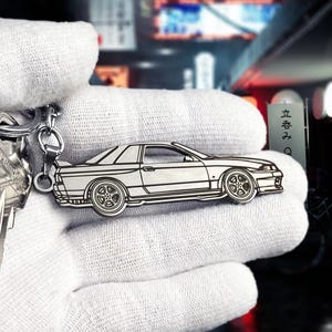 May include: Silver car-shaped keychain. The keychain features a detailed outline of a sports car with visible wheels, windows, and a spoiler. The car is attached to a metal chain and ring. The background is blurred.