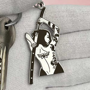 Puede incluir: Llavero plateado con una ilustración estilizada de una mujer sosteniendo una espada. La mujer tiene tatuajes en el brazo y un diseño de mariposa en el torso. El llavero está sujeto a un llavero.