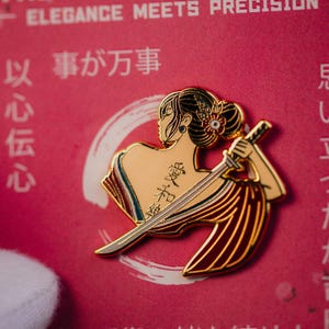 May include: A gold-colored enamel pin featuring a woman with a sword, with Japanese text. The pin has red, gold, and white accents. The background is a pink surface with Japanese text and the words "ELEGANCE MEETS PRECISION".