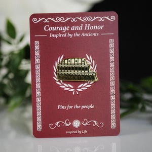 May include: A gold-colored Colosseum pin on a burgundy card with the text "Courage and Honor" and "Pins for the people". The pin is surrounded by a white laurel wreath. The card has decorative borders and the words "Inspired by Life".