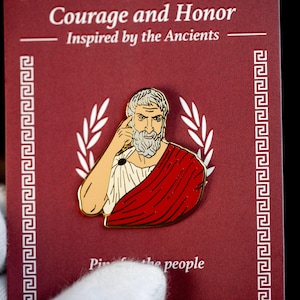 Pode incluir: Um alfinete dourado com um retrato de um homem barbudo com uma toga vermelha e uma túnica branca. O alfinete está em um cartão bordô com o texto "Courage and Honor" e "Inspired by the Ancients".