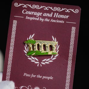 Op de afbeelding: Een goudkleurige pin met een gewelfde structuur met een boom, tegen een groen en beige landschap. De pin zit op een bordeauxrode kaart met de tekst "Courage and Honor" en "Inspired by the Ancients". De kaart luidt ook "Pins for the people".
