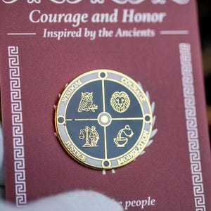 Puede incluir: Un pin redondo con borde dorado y centro gris oscuro, con símbolos de sabiduría, coraje, justicia y moderación. El pin está sobre un fondo burdeos con el texto "Courage and Honor" e "Inspired by the Ancients".