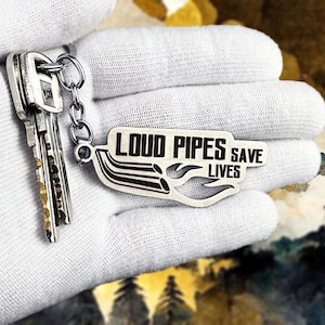 Peut inclure: Un porte-clés argenté avec le texte "LOUD PIPES SAVE LIVES" en noir. Le porte-clés présente un design stylisé de tuyau d'échappement et est attaché à un jeu de clés.