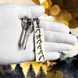 Könnte beinhalten: Ein Schlüsselbund mit einem silberfarbenen Metallschlüsselanhänger. Der rechteckige Schlüsselanhänger hat japanische Schriftzeichen eingraviert. Die Schlüssel und der Schlüsselanhänger werden in einer weißen behandschuhten Hand gehalten.