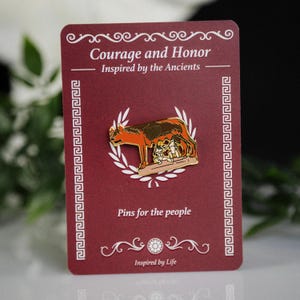 May include: A red card with the text "Courage and Honor" and "Inspired by the Ancients." A gold-colored pin depicts a wolf nursing two babies, surrounded by a laurel wreath. The text "Pins for the people" and "Inspired by Life" are also visible.