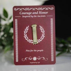 Puede incluir: Un pin dorado con forma de columna clásica, sobre una tarjeta burdeos. La tarjeta presenta el texto "Courage and Honor" y "Pins for the people". Una corona de laurel blanca rodea el pin, con bordes decorativos.